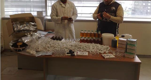 L’immagine mostra due persone in un laboratorio, con una grande quantità di oggetti disposti su un tavolo. Tra questi: Numerose siringhe (vuote o usate) sparse sulla superficie. Bottiglie di vetro con liquidi di colore arancione/ambra, probabilmente medicinali o sostanze chimiche. Scatole di medicinali impilate, alcune con etichette visibili. Un contenitore di plastica bianco, forse per rifiuti o materiali. Un oggetto nero con un simbolo giallo (simile a un simbolo di radiazione), che indicare materiali pericolosi o radioattivi.
Una persona indossa un grembiule bianco da laboratorio, l’altra un giubbotto con la scritta “N.A.S.”, suggerendo un’operazione di sequestro.
